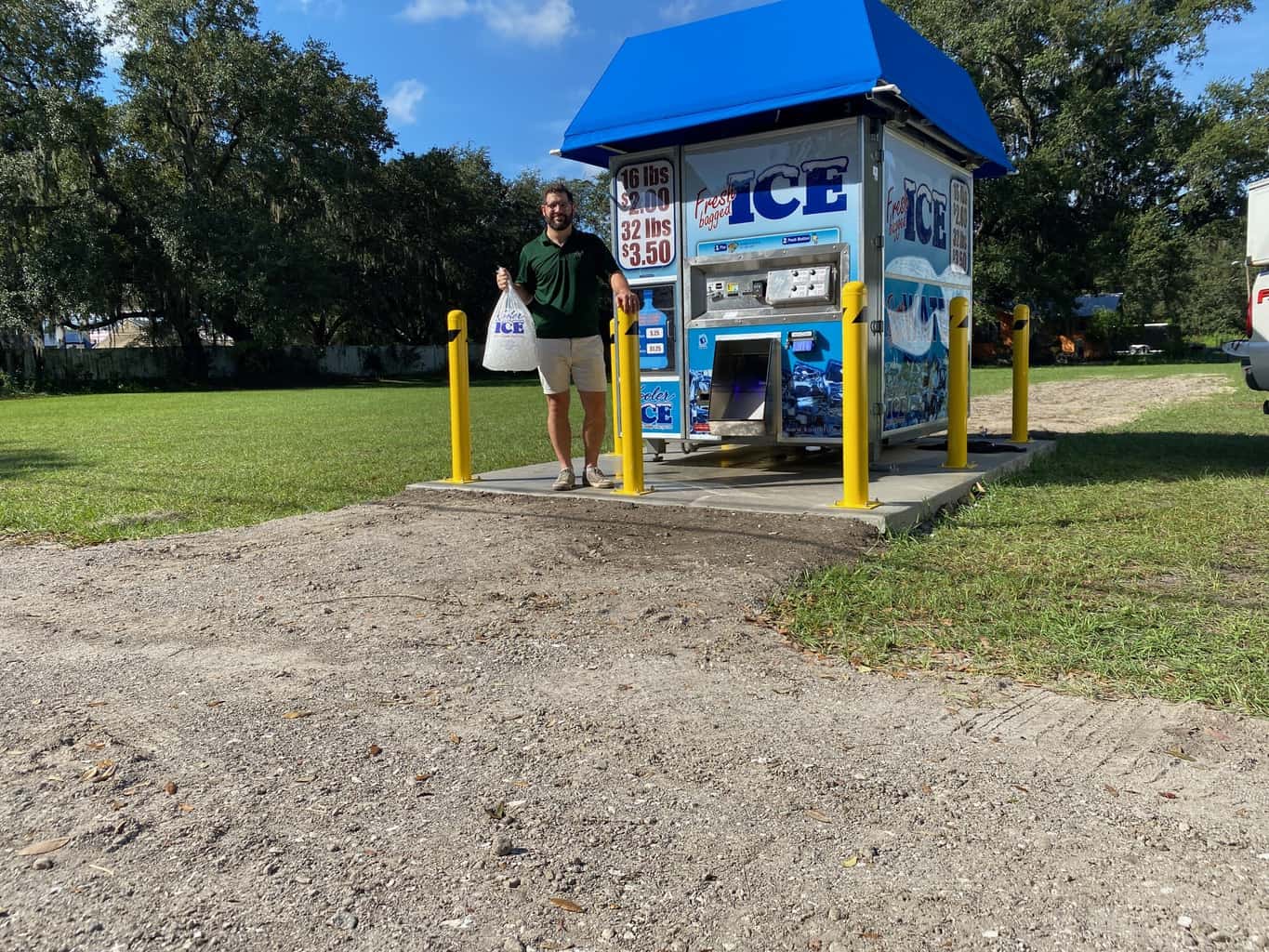Man stands with a bag of ice beside a blue-roofed ice vending kiosk in a grassy park, surrounded by yellow bollards.