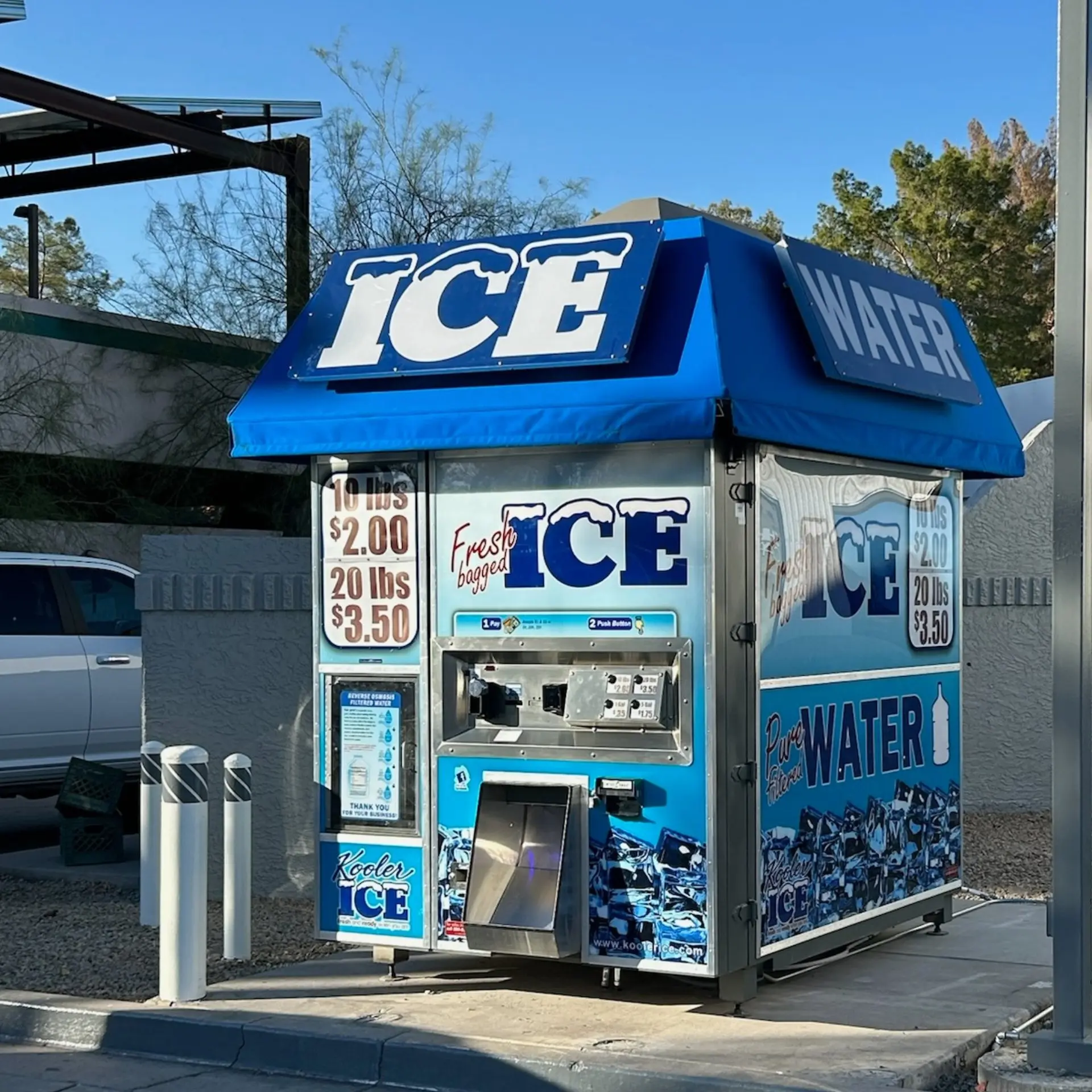 Outdoor ice and water vending kiosk with bold ICE and WATER signs on a blue canopy; pricing panels are visible on the side.